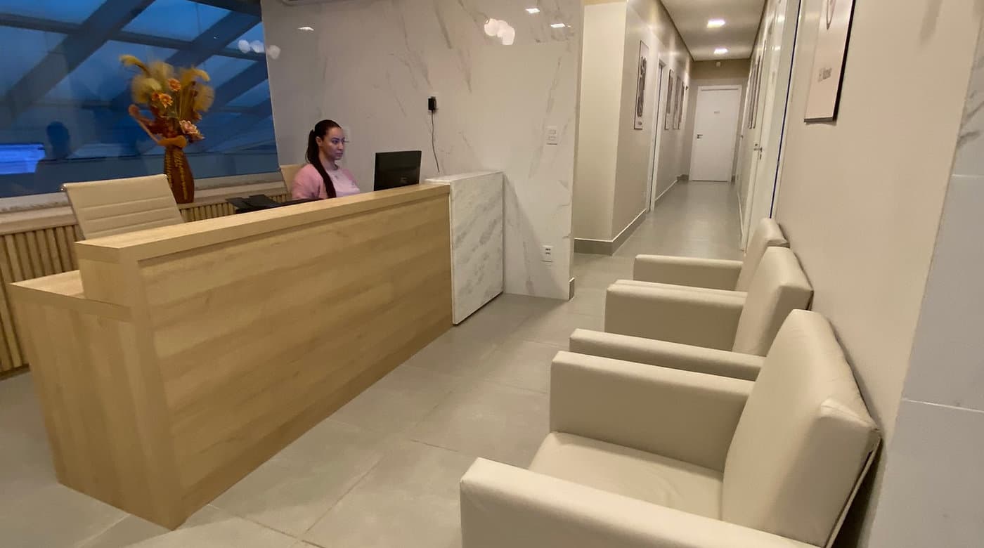 Recepção da unidade Lourdes do Instituto Hiraki, em Belo Horizonte: balcão de madeira, recepcionista e poltronas de espera